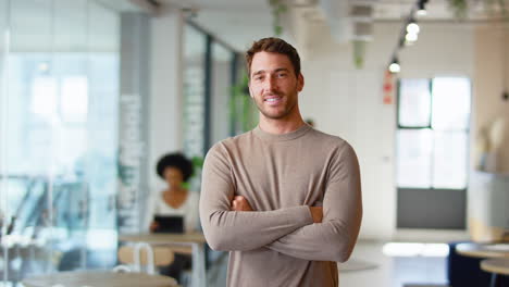 Portrait-Of-Smiling-Businessman-Standing-In-Busy-Modern-Open-Plan-Office