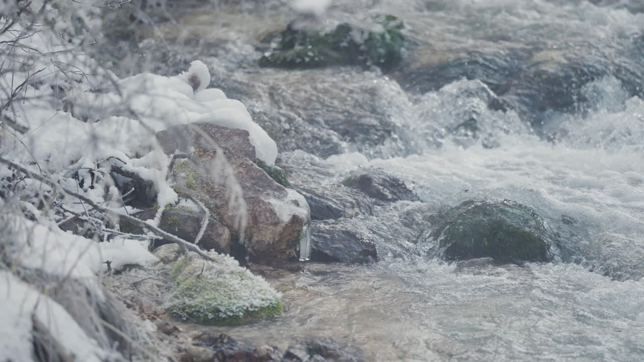 A shallow mountain stream flows in the rocky riverbed surrounded by the snow-covered low banks and frozen branches. Parallax video