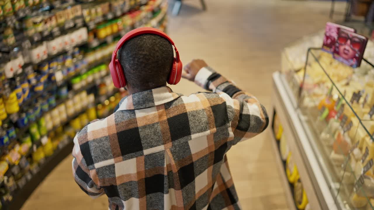 Top view of a man with Black skin color with a short haircut in a checkered shirt and in red wireless headphones dances and walks with a cart between the counters in a modern grocery store