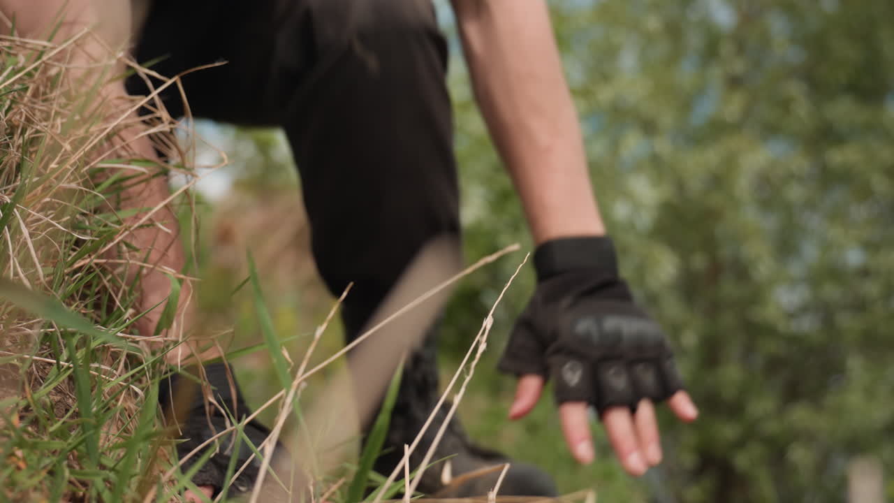 young man emerging from grass hiding spot, hand on ground, brushing soil from cargo pant leg, blurred grass foreground, tank top upper body, bright sky backdrop, rugged survival with tactical mood
