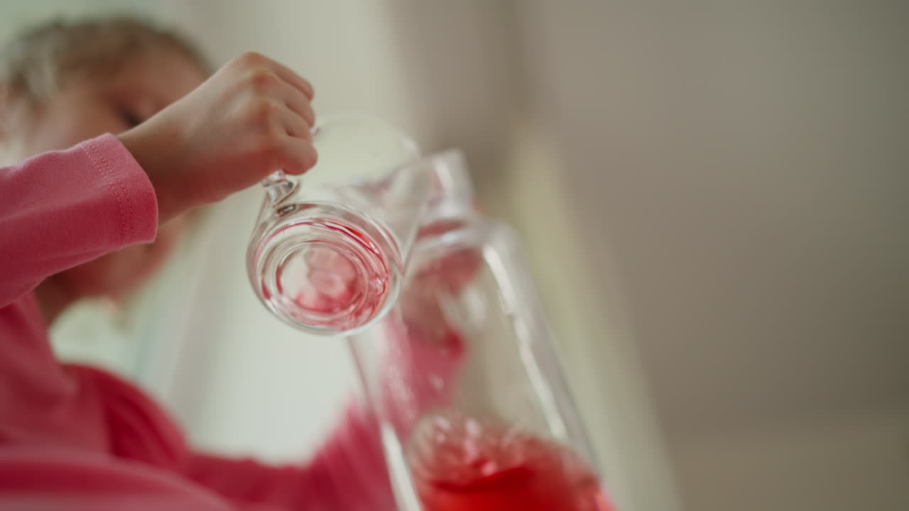 Low angle of grand daughter seated by window in kitchen drinking red juice from glass jar, accidental drip onto pink dress, small hands gripping handle, soft daylight and blurred interior background
