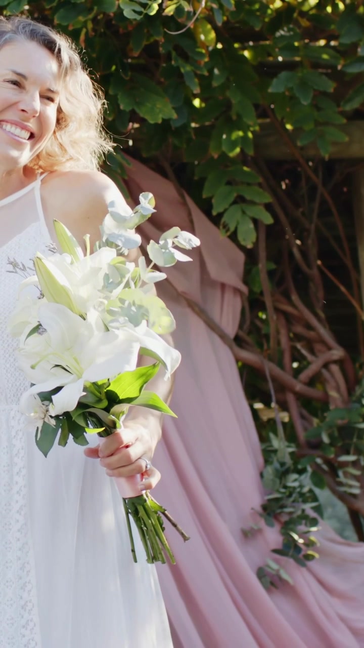 Video verticale: Sposa che entra da sinistra in abito bianco, tiene un bouquet in giardino, con gli invitati che applaudono.