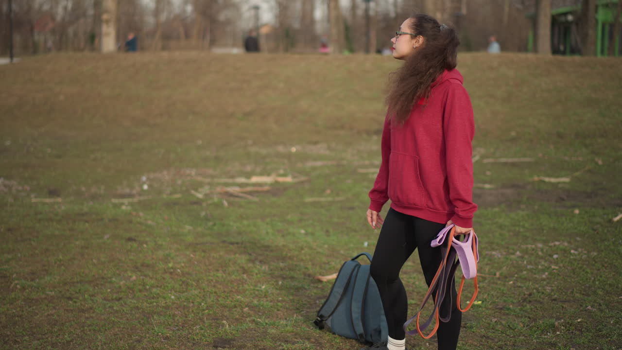 Asian Woman Walking In Park Backpack Leash In Hand, Slow Departing Stride Across Muddy Grass Toward Tree Line, Casual Hoodie, Ponytail Swinging, Overcast Light, Contemplative Mood And Solitary Outdoor