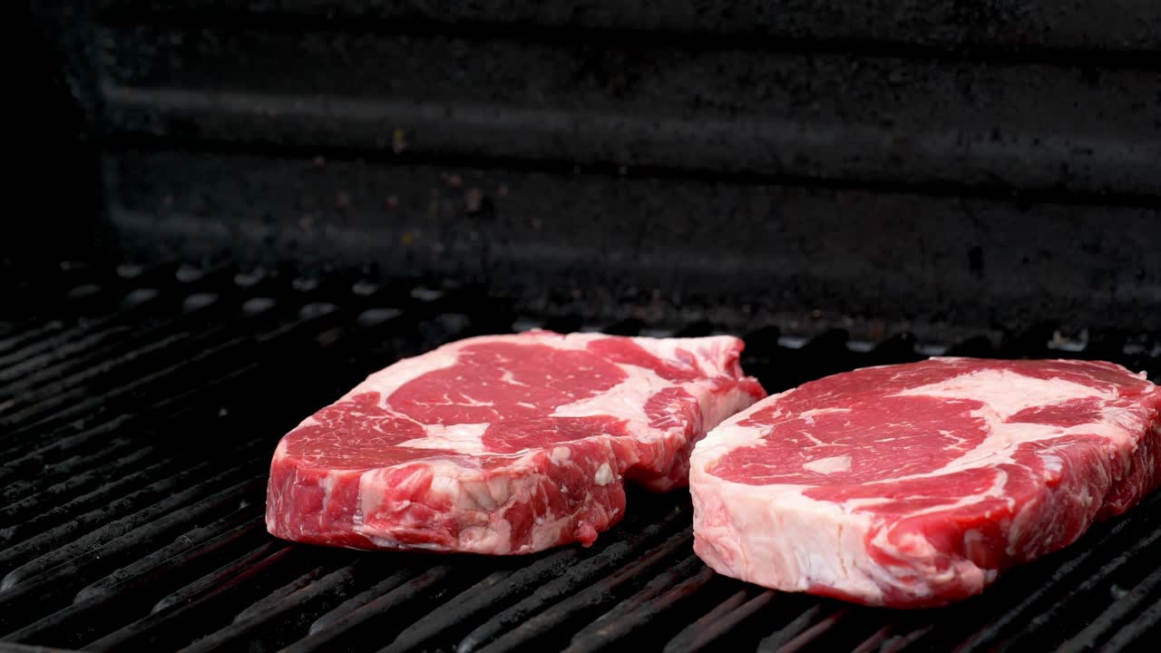 Two raw rib-eye steaks getting ready to cook on a grill