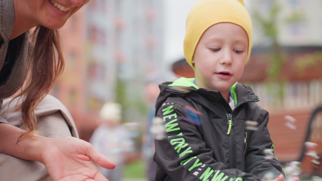 Close up of boy in yellow beanie playing with soap bubbles outdoors, focused expression as child swipes hands through floating spheres while parent nearby