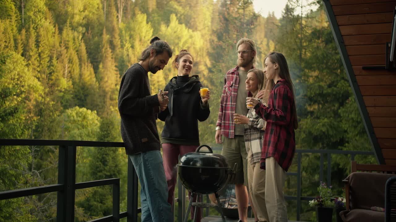 The guy plays the guitar, and his friends with yellow drinks in their hands dance during a picnic on the spacious balcony of a country house overlooking the mountains and coniferous forest