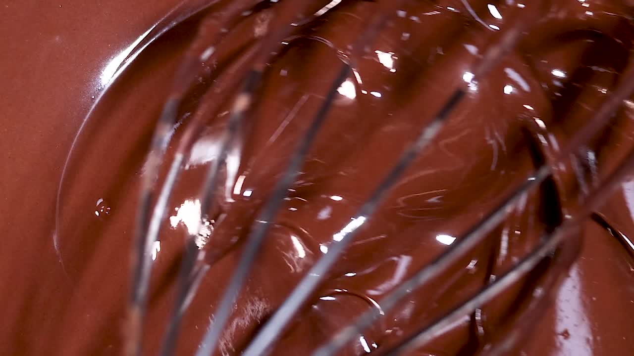A whisk gently stirs a smooth, glossy chocolate mixture, capturing its rich texture and shine.