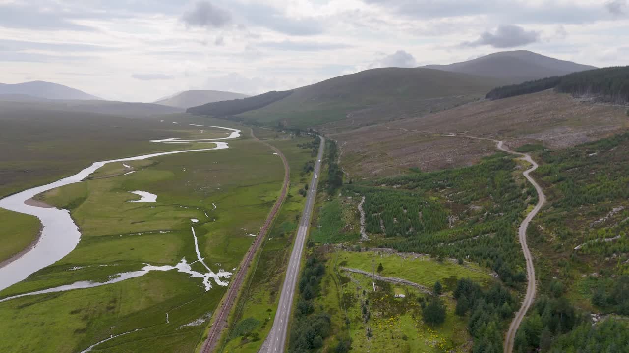 Drone footage glides above a winding road, river, and patchwork of deforested and green terrain in the Scottish Highlands under overcast daylight