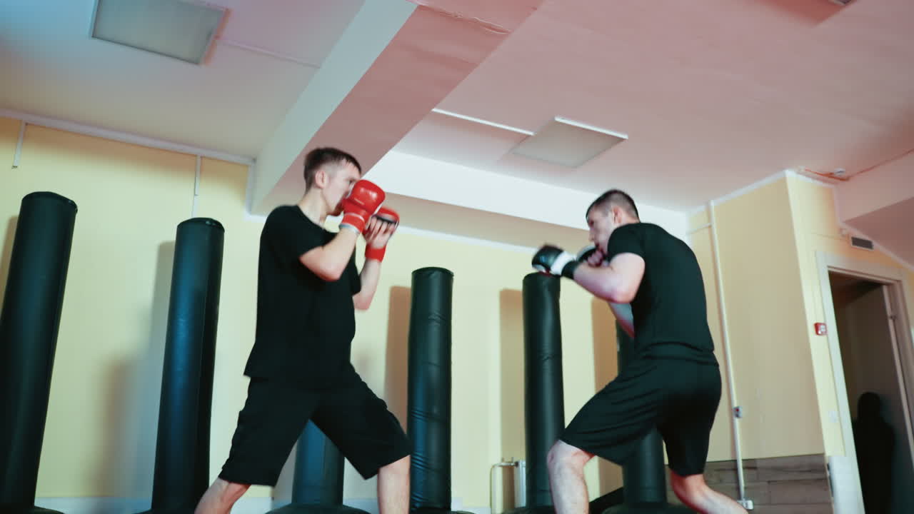Wrestlers sparring inside gym, wearing gloves and black outfits, facing each other in combat stance while preparing for martial training on red mat floor surrounded by standing punching bags