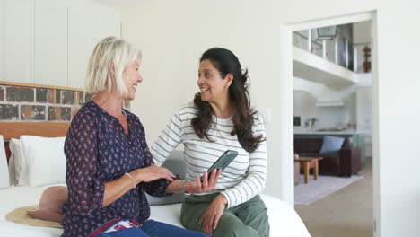 Two-happy-diverse-senior-women-using-smartphone-and-laughing-in-sunny-bedroom,-slow-motion