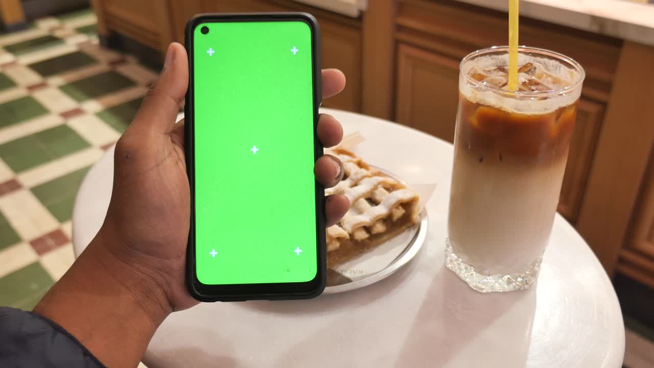 Phone with green screen display in hand, latte, and apple pie at a cafe