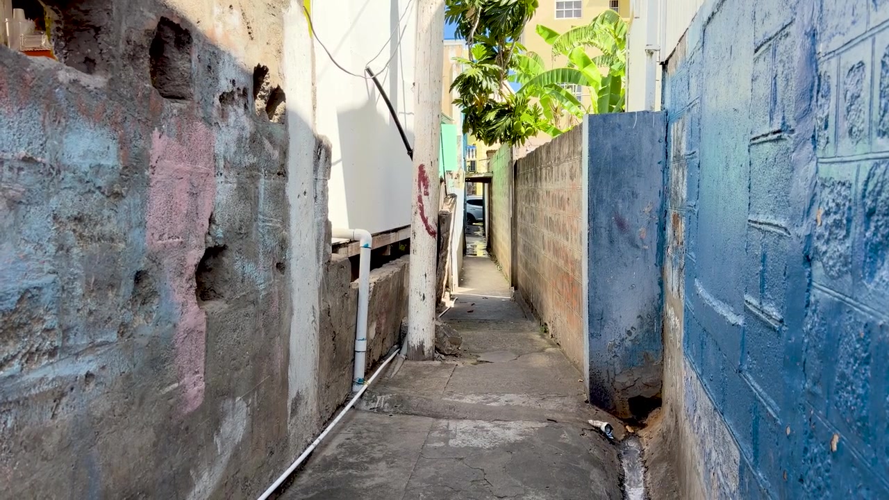 View down a narrow, empty urban alleyway featuring weathered concrete walls, peeling blue paint, and a pathway leading to a sunlit street in a Caribbean town.