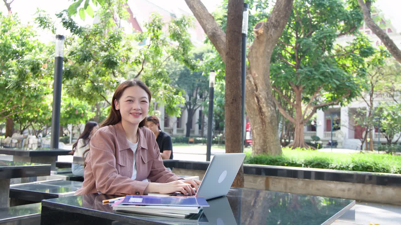 一位年輕亞洲女性在戶外校園的桌子旁，用手提電腦和教科書學習。