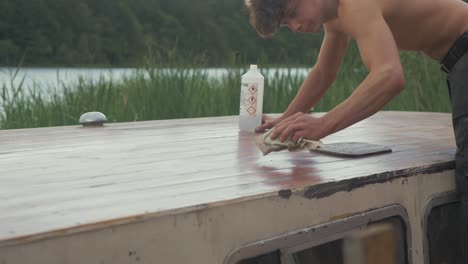 young carpenter brushing down sanded roof planking of old wood boat using acetone on cloth in hot summer heat