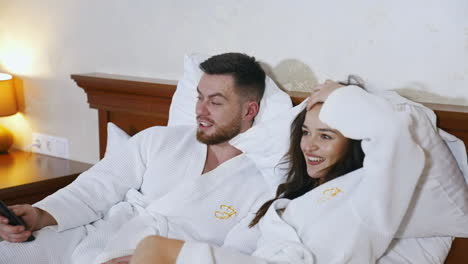 lovely couple on bed in hotel. attractive young woman and handsome man in white coats lying in bed and watching movie on tv together.
