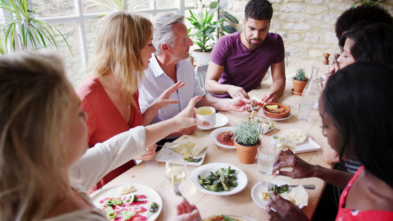 En flerkulturell gruppe voksne venner i forskjellige aldre spiser tapas sammen til lunsj på en liten restaurant, sett ovenfra.