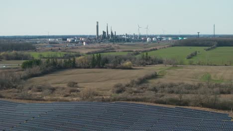 Amplia-Vista-De-Paneles-Solares-Con-Telón-De-Fondo-Industrial-Y-Turbinas-Eólicas,-Campos-Verdes-Vibrantes-Rodean-La-Zona,-Durante-El-Día,-Soleado