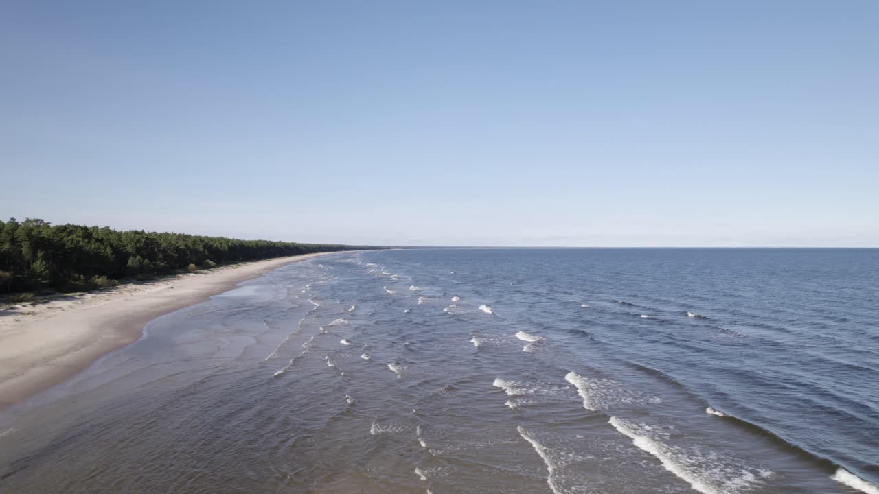 Oddalające się ujęcie z drona: długie, równoległe fale rozbijające się na piaszczystych płyciznach bałtyckiego wybrzeża.