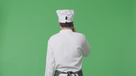 vista trasera del chef asiático hablando en el teléfono inteligente mientras camina en el estudio de fondo de la pantalla verde