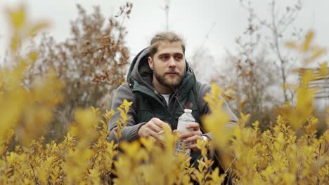 hombre fumando en un campo con flores amarillas mientras sostiene una botella de agua