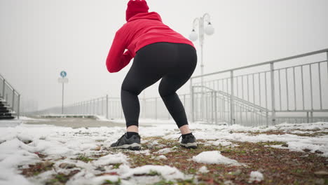 lady performing jump squats during winter outdoor workout on snowy ground, wearing red hoodie and black gloves, background includes light poles, iron railing, signpost, and foggy atmosphere