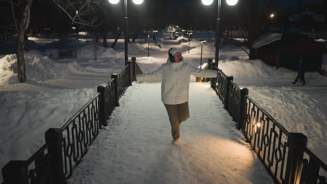 Elegant woman in white puffer coat and headphones dances gracefully on snow covered walkway under glowing lampposts and string lights against dark winter sky with flowing expressive gestures
