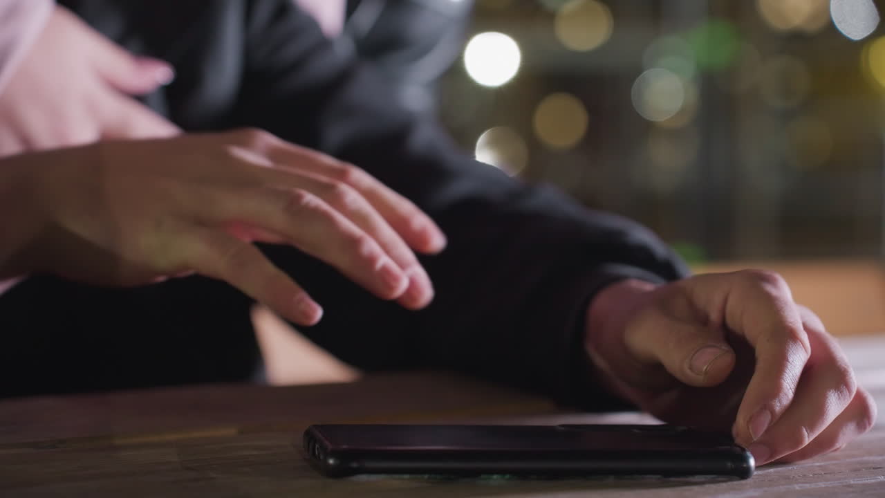 Close up hand view of person holding phone on wooden table as another hand gently places around him and phone slips down onto table with warm bokeh lighting in soft urban night background