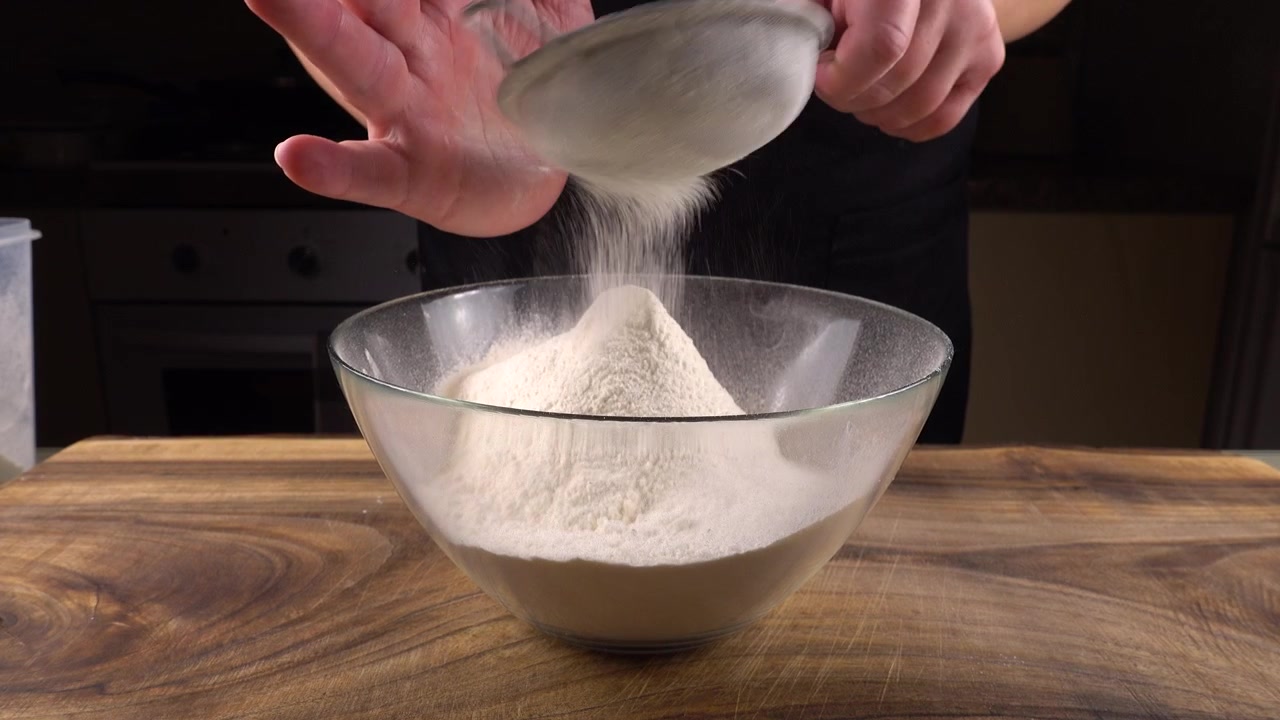 Mengayak tepung menggunakan saringan ke dalam mangkuk kaca di dapur.