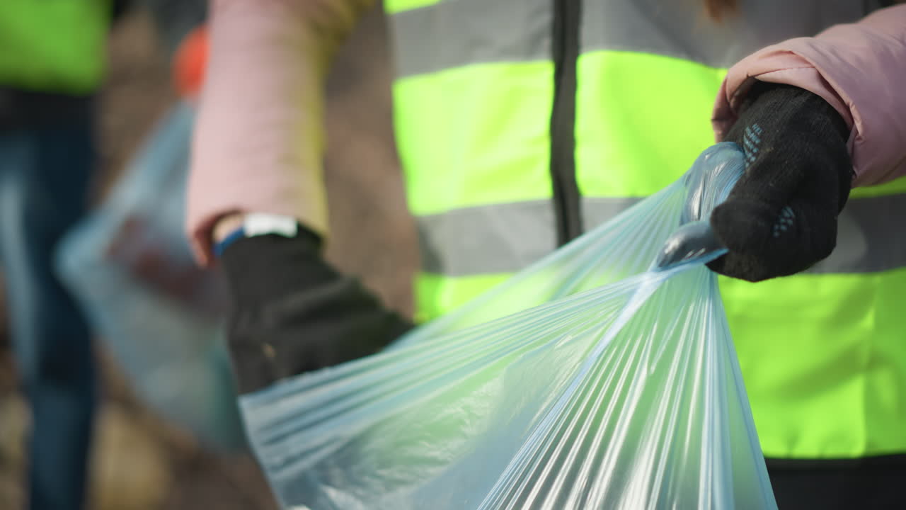 Volunteer wearing black gloves picking up dirty glass bottle from ground covered with dry leaves and branches during outdoor cleanup, placing litter into blue plastic bag to protect nature