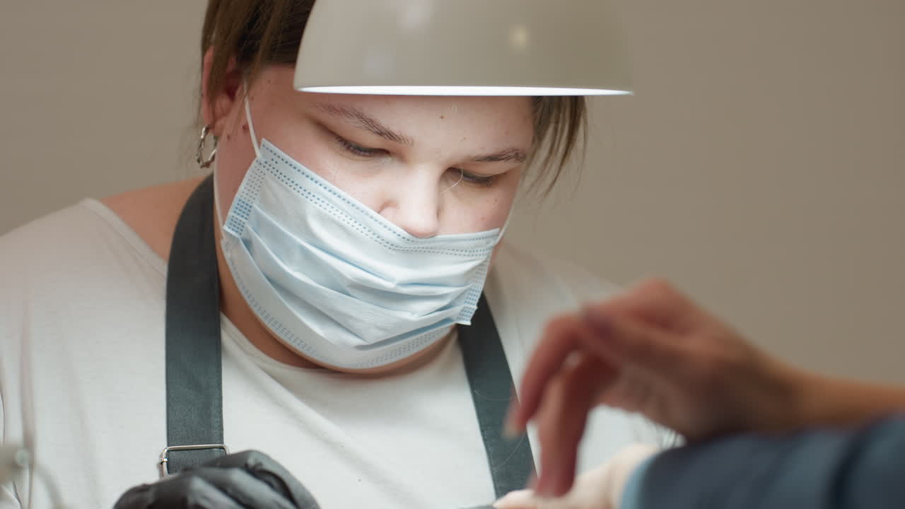 Nail technician in white shirt and dark apron focused on task while wearing face mask, illuminated by overhead light, attending to client with careful precision in professional salon environment