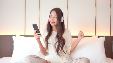 portrait of beautiful exotic woman listening music from smartphone with wireless headphones in bed, full frame slow motion