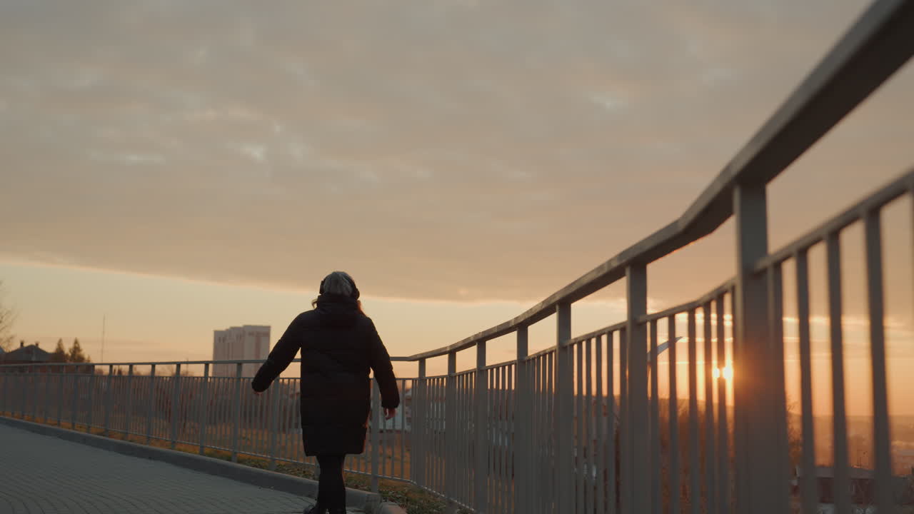 Baile en puente al atardecer, mujer asiática en silueta, movimiento lento y expresivo en un paseo ribereño, abrigo grueso y gorro de punto, cálida luz dorada, líneas de la barandilla, estado de ánimo reflexivo y coreografía tranquila.