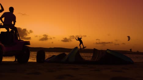 Slomo-Aufnahme-In-Der-Dämmerung-Eines-Kitesurfers,-Der-Vor-Freunden-Springt,-Die-Auf-Einem-Buggy-Sitzen---Dunkle-Silhouetten-Vor-Orangefarbenem-Sonnenuntergangshimmel