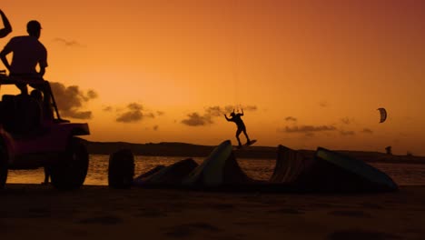 slomo shot at twilight of kitesurfer jumping in front of friends sitting on buggy - dark silhouettes against orange sunset sky