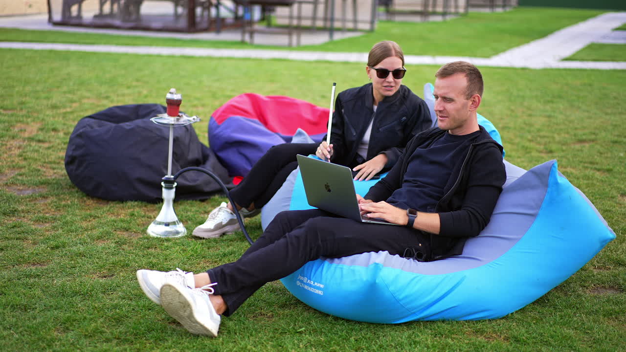 Man and woman communicating spending time outdoors. Man is busy with his work on laptop and lady smoking hookah and speaking. Blurred backdrop.