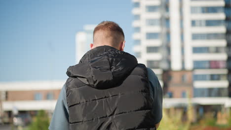 back view of white boy in black puffy vest walking along path in modern city area with tall residential buildings under bright sky during sunny day in autumn