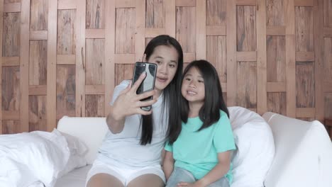 4k asiática sonriendo mujeres felices con su hija usan teléfono inteligente para video llamada en línea en la habitación del hotel. familia feliz llamando video chat en teléfono móvil en el hotel. mujer sentada en la cama y haciendo video llamada en liña en la villa del hotel