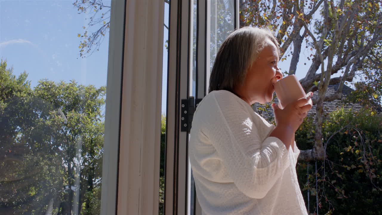 Drinking coffee, senior woman enjoying morning sunlight by open window, copy space