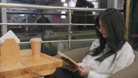 young lady with long dark hair and glasses sits comfortably in restaurant flipping through book, hand with floral tattoo resting near coffee cup on wooden table, evening ambiance in background