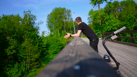 tattooed man with black clothing walking on a bridge up to the wooden railing and watching around while thinking and observing the nature around him