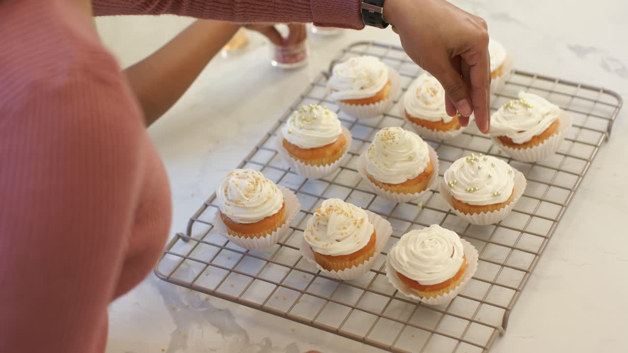 Mãe e filha diversas em casa, polvilhando granulado rosa e dourado em cupcakes, com espaço para texto