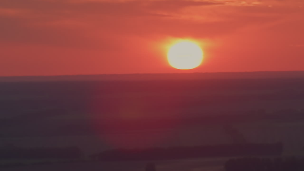Blazing orange sun setting over distant horizon bathes evening sky and rolling landscape in warm glow while subtle flare blooms around bright solar disc capturing tranquil dusk