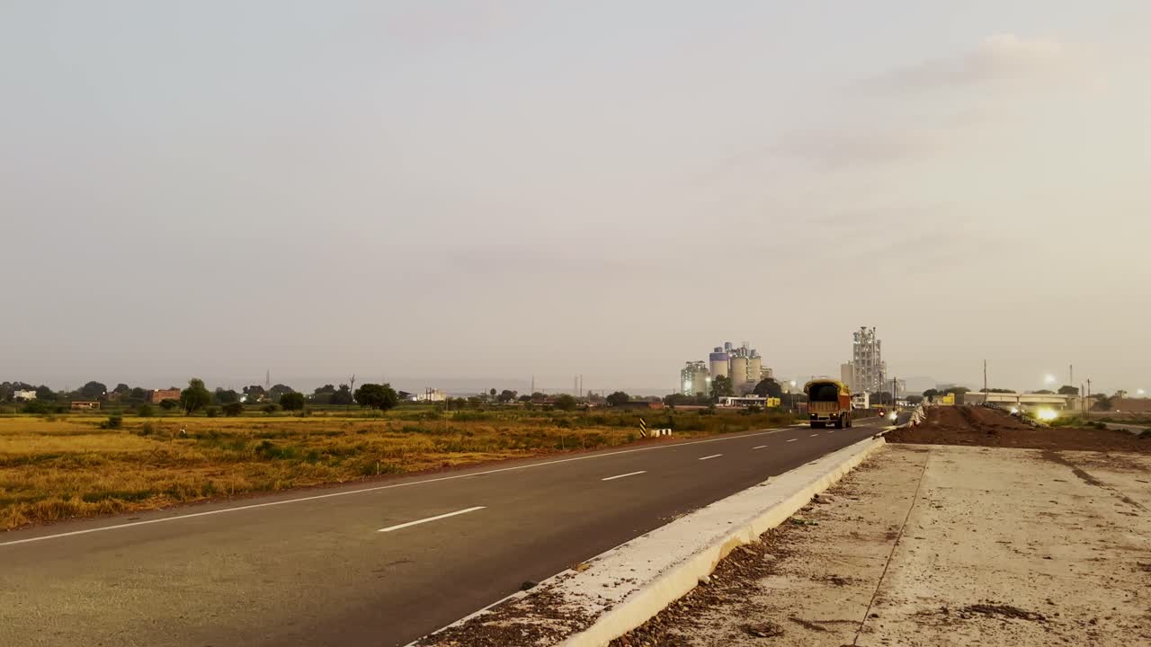 Static shot capturing a transitional landscape, blending rural elements with industrial development under a muted sky, as a paved road stretches across the mid-ground, leading towards cement factory