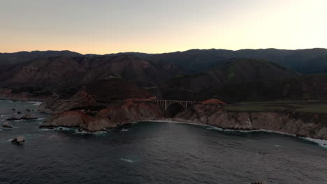 California-coast-with-Bixby-Bridge-in-Big-Sur-at-sunrise,-drone-backwards