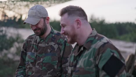 soldiers in camouflage clothing, discussing in the evening at a common bonfire frying sausages for supper, men joking, laughing, resting after work, service, field exercises