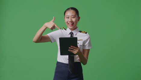 asian woman pilot smiling and pointing to tablet in her hand while standing in the green screen background studio