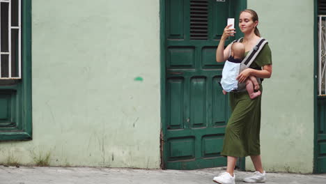 Una-Mujer-Joven-Viaja-Con-Su-Bebé-En-Una-Mochila-Canguro,-Capturando-Momentos-En-Su-Teléfono-Móvil.-Camina-Tranquilamente,-Mirando-De-Vez-En-Cuando-La-Pantalla-Del-Teléfono