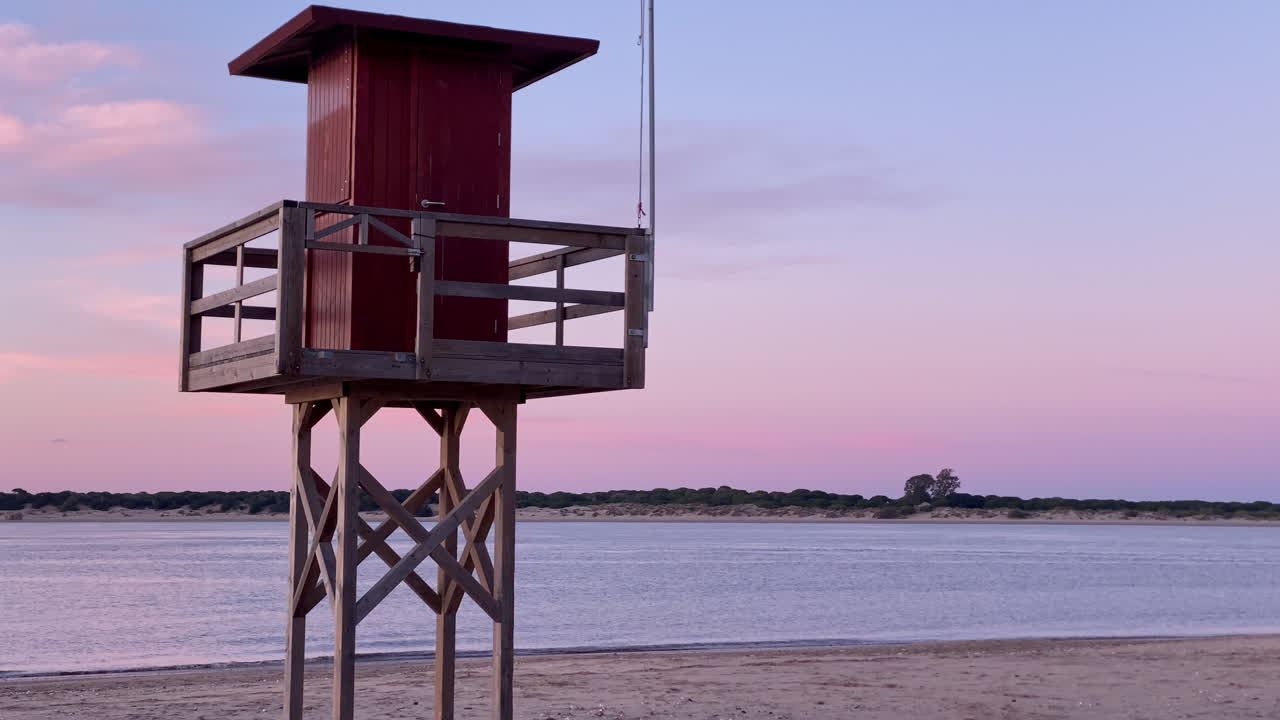Livreddertårn på en sandstrand ved solnedgang, der viser en gradueret himmel med en flodmunding og både i baggrunden.