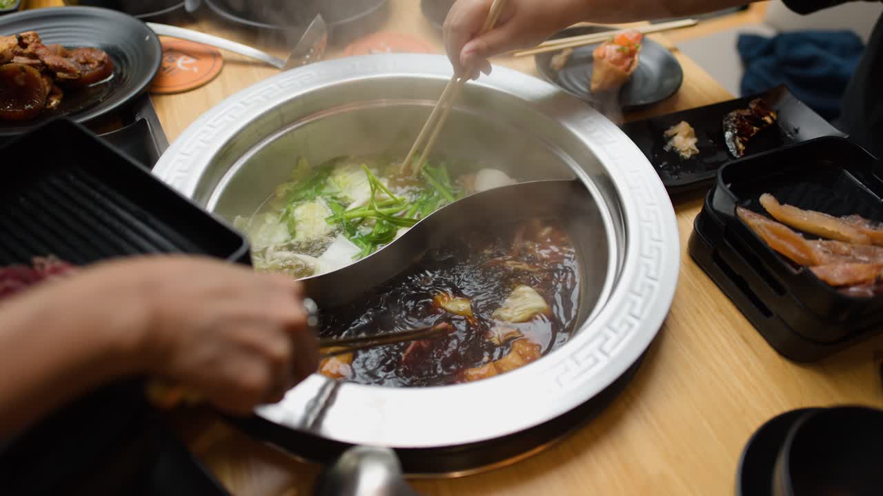 Hand dips sliced meat into steaming dual broth hotpot, surrounded by vegetables and side dishes