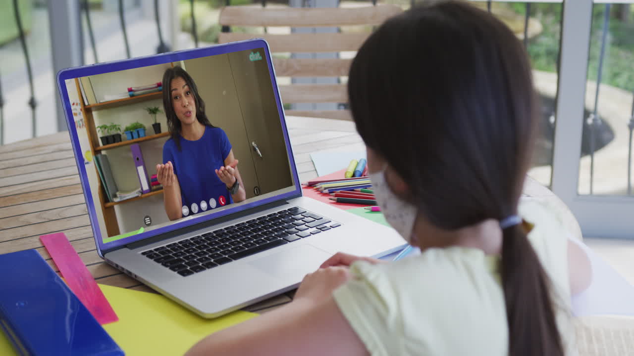 vista posterior de una niña con una máscara facial haciendo una videollamada con una maestra en una computadora portátil en casa